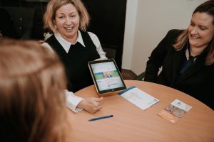 Three people sitting at a round table. One woman is pointing to something on an electronic tablet.