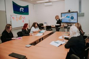 two people sitting at a desk looking at another person pointing at a big screen as if in a training workshop