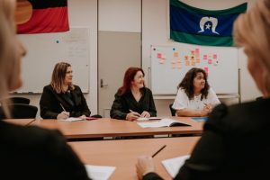 Three people sitting at a desk looking towards the front of the room. In the background is an Aboriginal flag and a Torres Strait Islander Flag, and a whiteboard with post-it notes