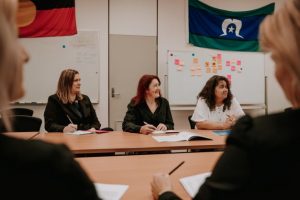 Three people sitting at a desk looking towards the front of the room. In the background is an Aboriginal flag and a Torres Strait Islander Flag, and a whiteboard with post-it notes